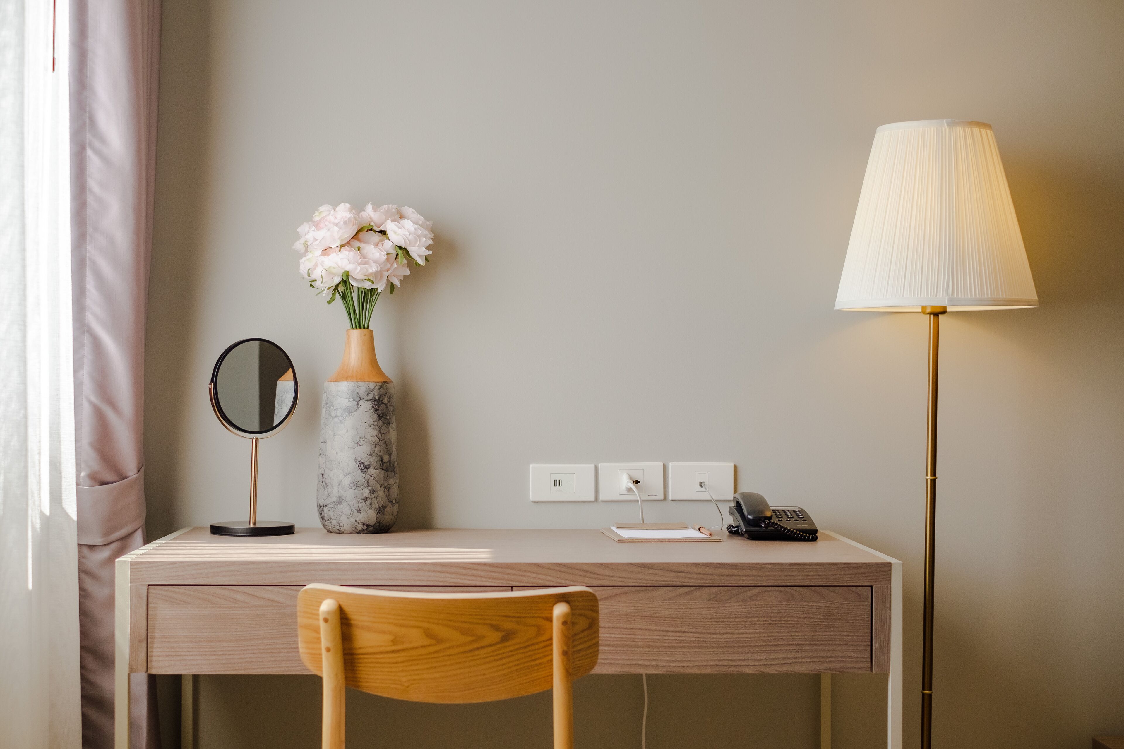 A neatly arranged desk with a vase of flowers, a mirror, a lamp, and a phone.