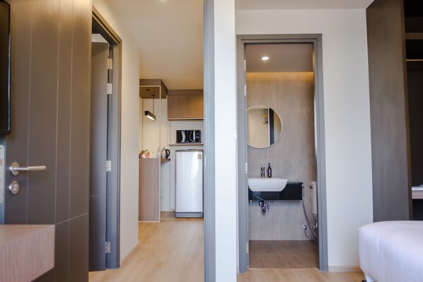 Una habitación de hotel moderna con piso de madera, una cama, un baño con lavamanos y espejo, y una kitchenette con refrigerador y cafetera.