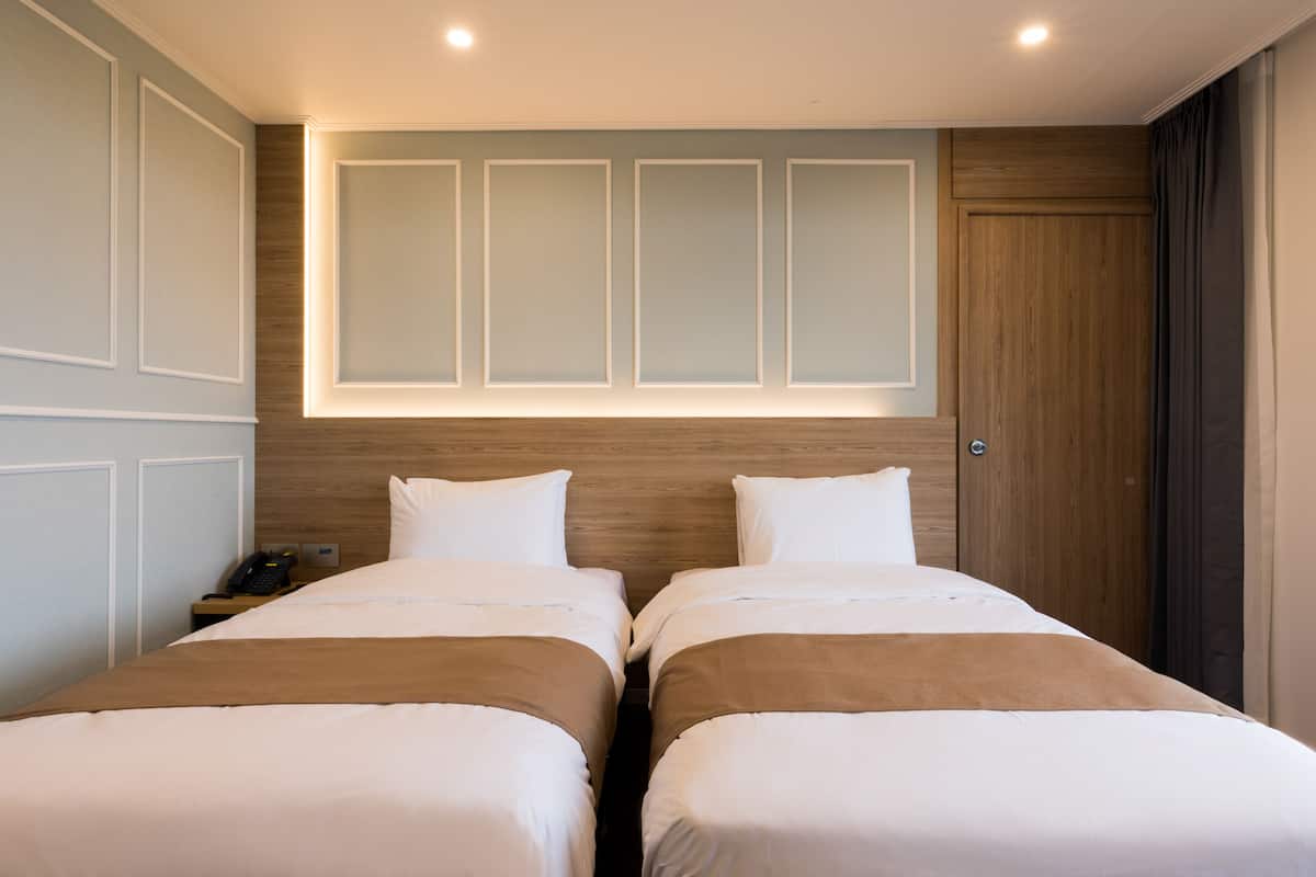 A hotel room with two beds, a wooden headboard, a nightstand with a phone, and a wall-mounted light fixture.