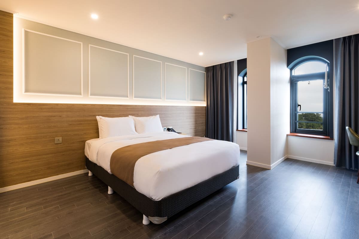 A modern hotel room with a large bed, wooden paneling on the walls, a view of the outdoors through a window, and a built-in headboard.