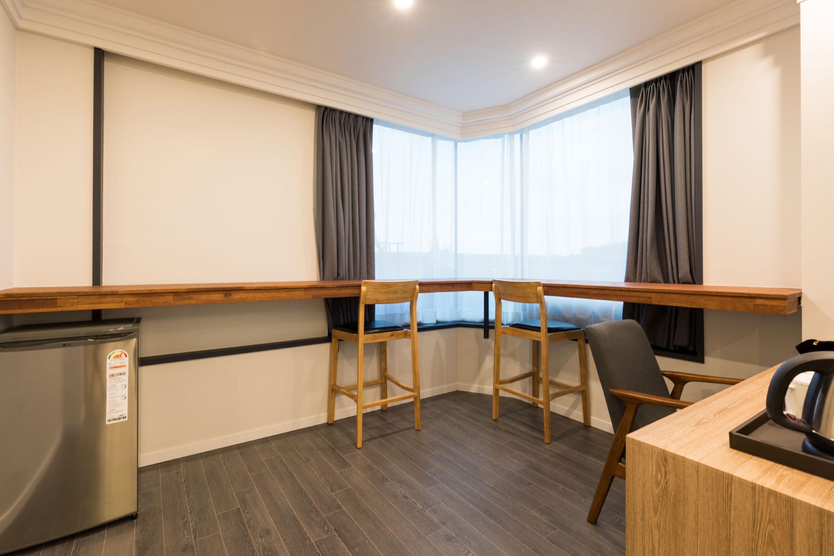 A hotel room with a wooden desk, two chairs, a small refrigerator, and a large window with curtains.