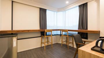 A hotel room with a wooden desk, two chairs, a small refrigerator, and a large window with curtains.