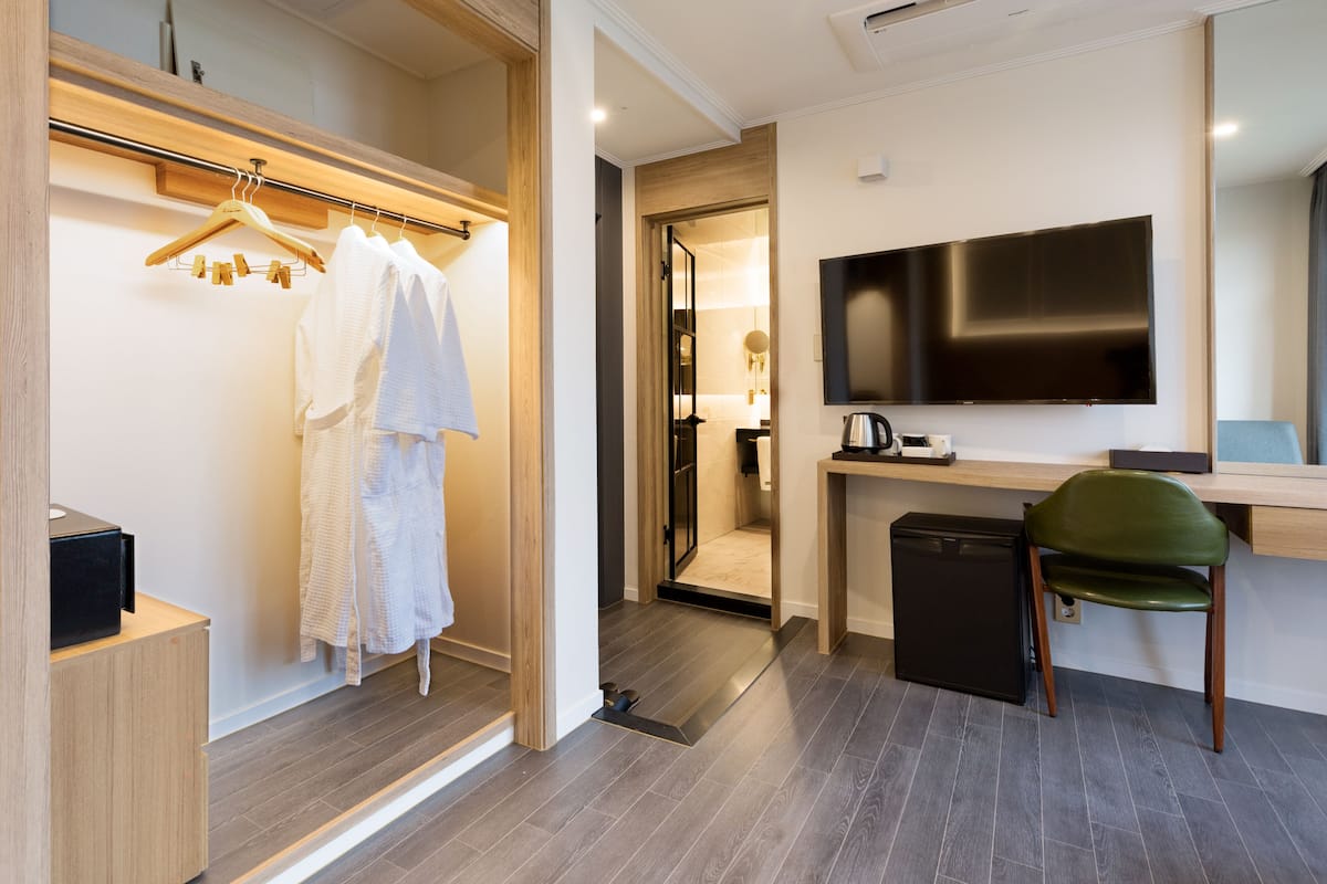 A modern hotel room with a walk-in closet, a desk with a chair, a television, and a small kitchenette.