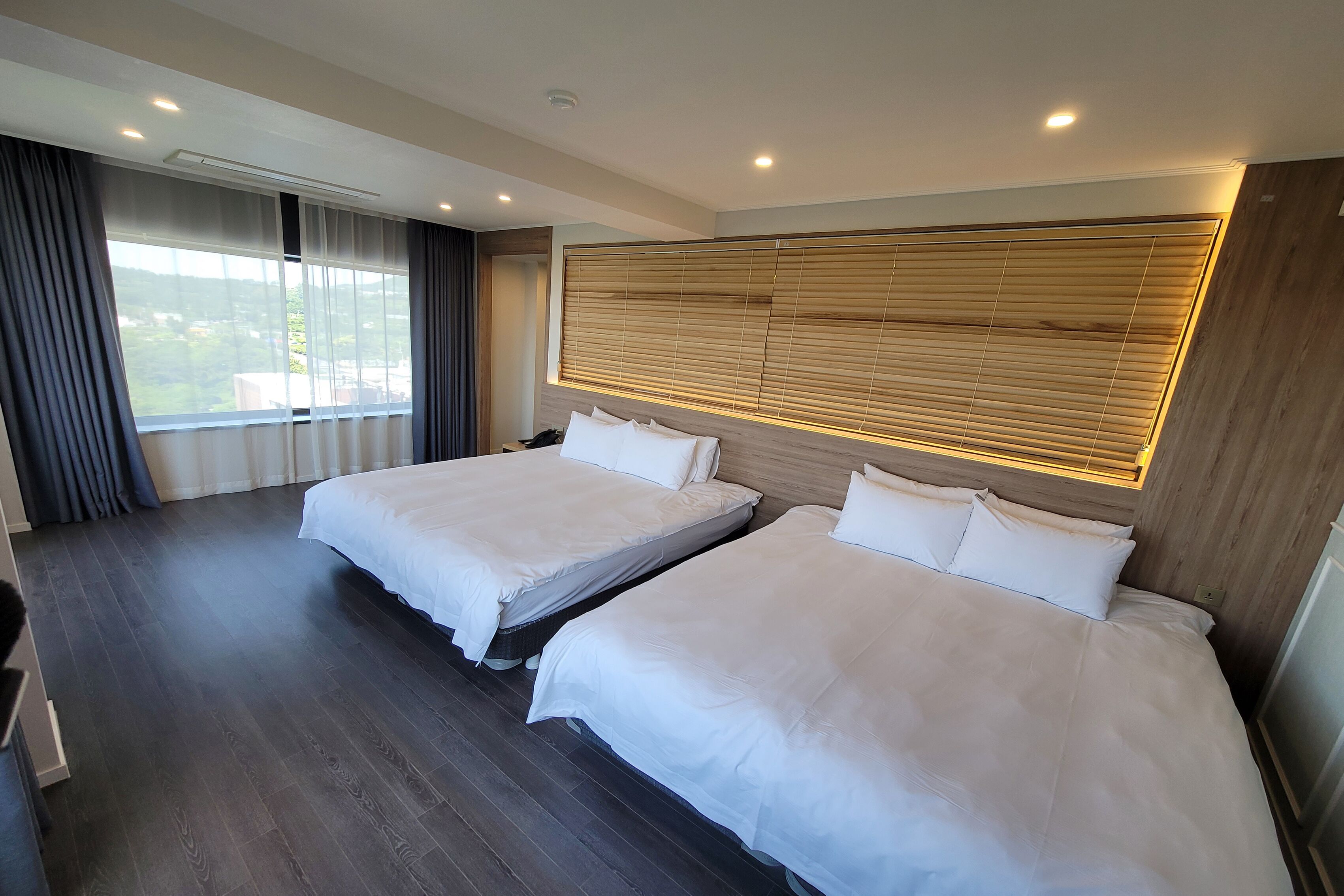 A hotel room with two beds, a large window with curtains, a wooden headboard, and a modern ceiling design.