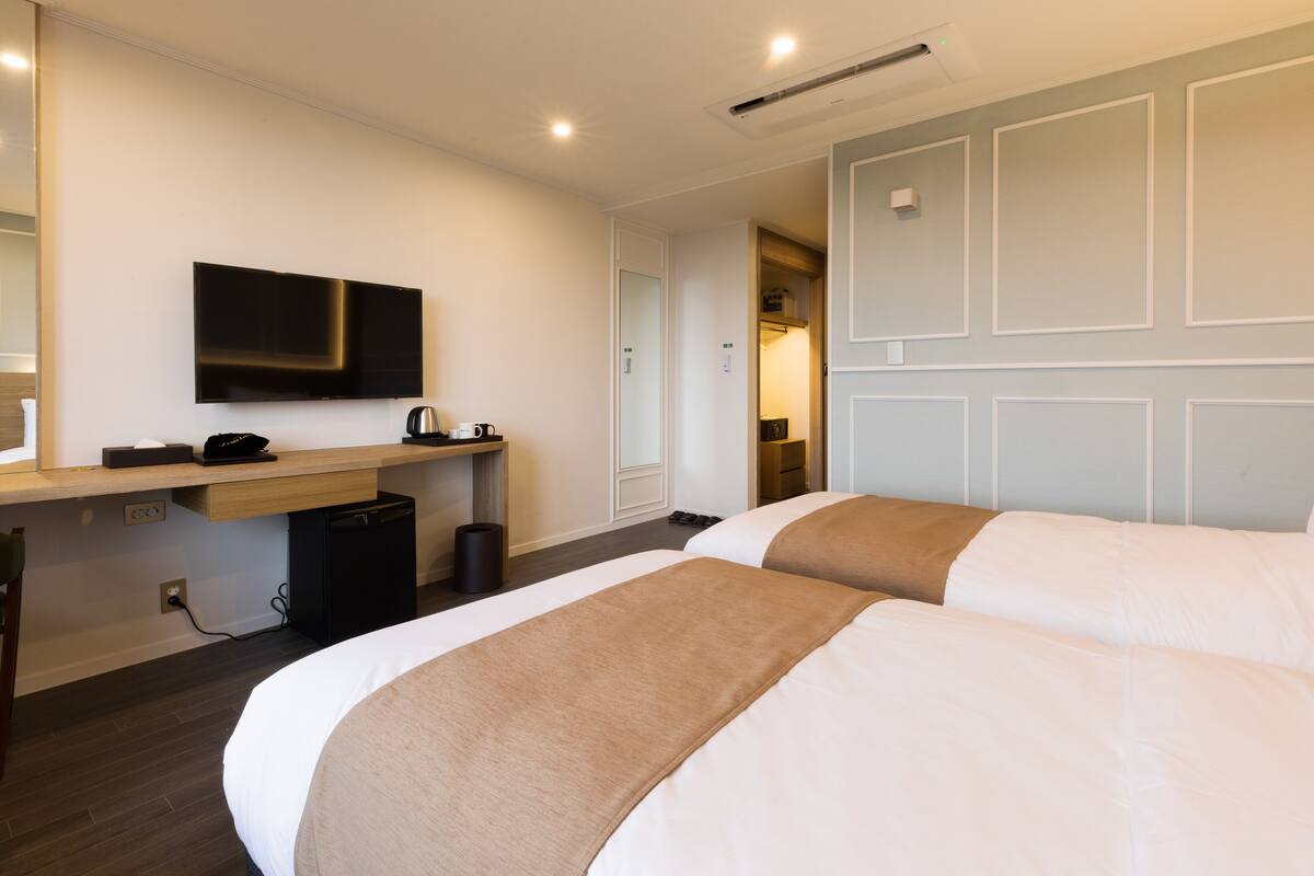 A hotel room with two beds, a desk with a flat-screen TV, a coffee maker, and a trash bin.