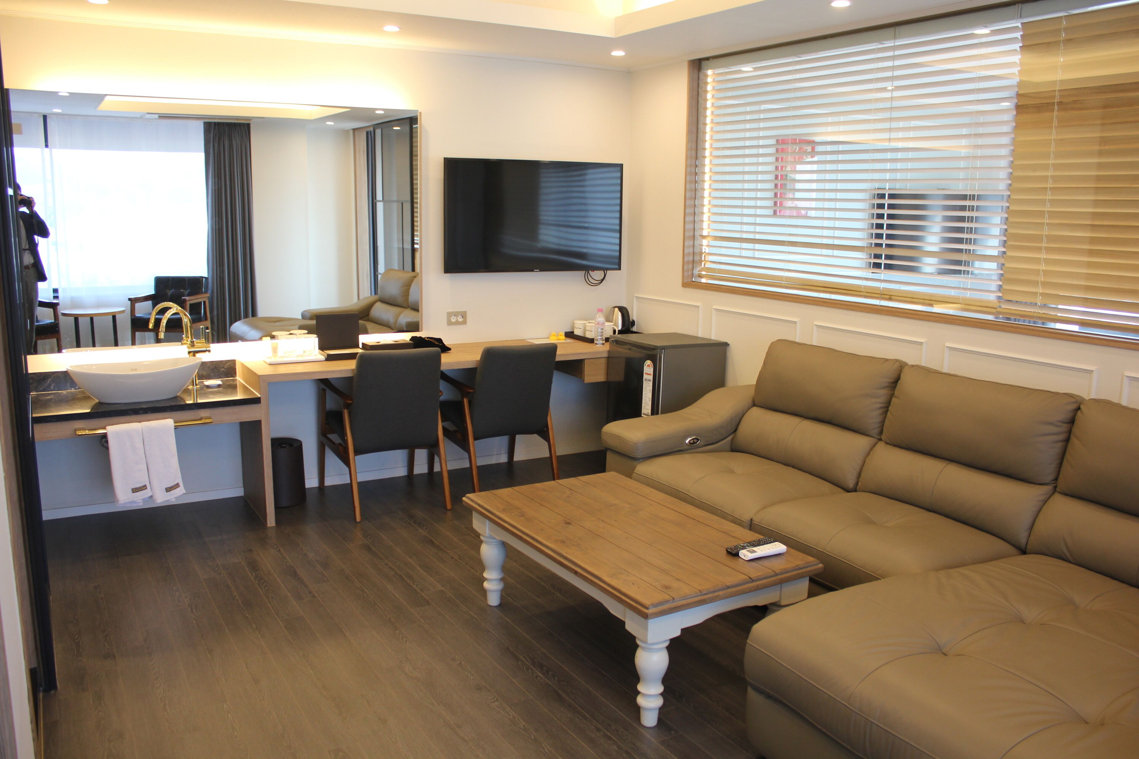 A modern hotel room with a wooden dining table, a sofa, a TV mounted on the wall, and a large window with blinds.