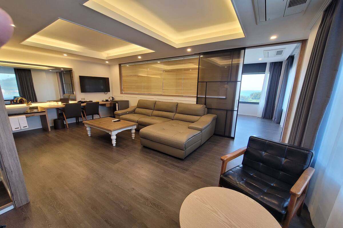 A modern living room with a large sofa, a coffee table, and a TV. There is a kitchen area with a dining table and chairs. The room has a large window with blinds.