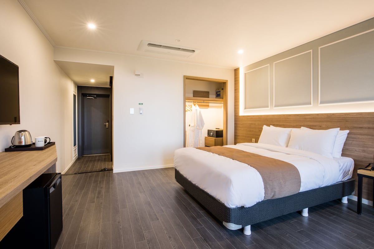 A modern hotel room with a large bed, a wooden bedside table, a built-in wardrobe, and a small kitchenette area.