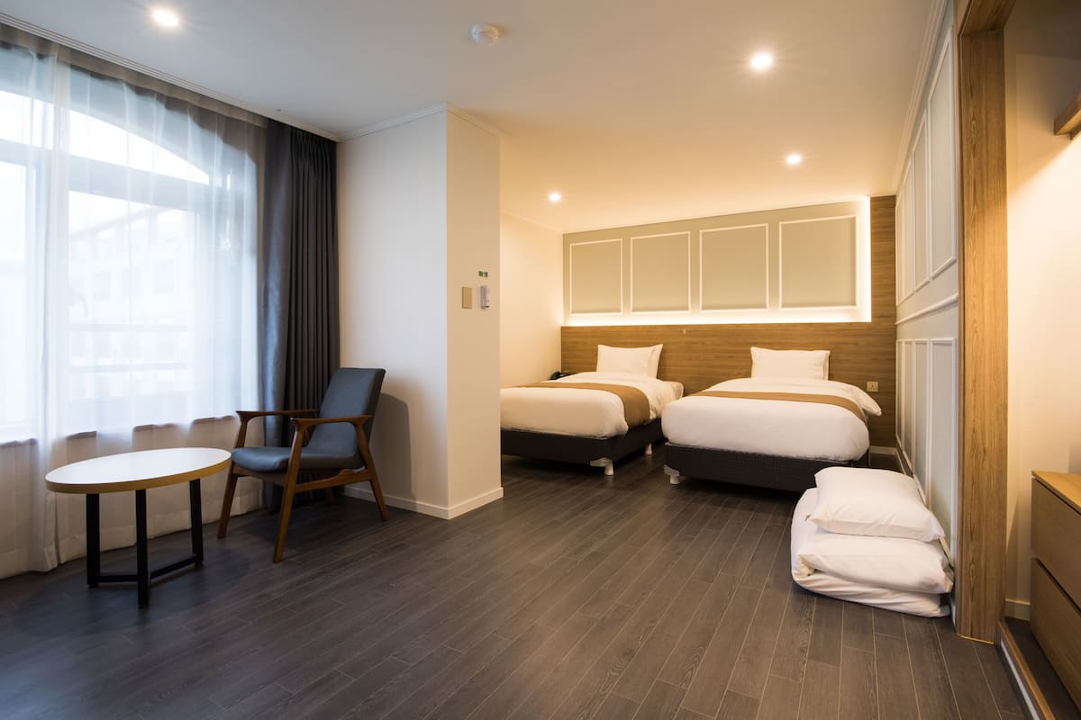 A hotel room with two beds, a small table, a chair, and a large window with curtains.
