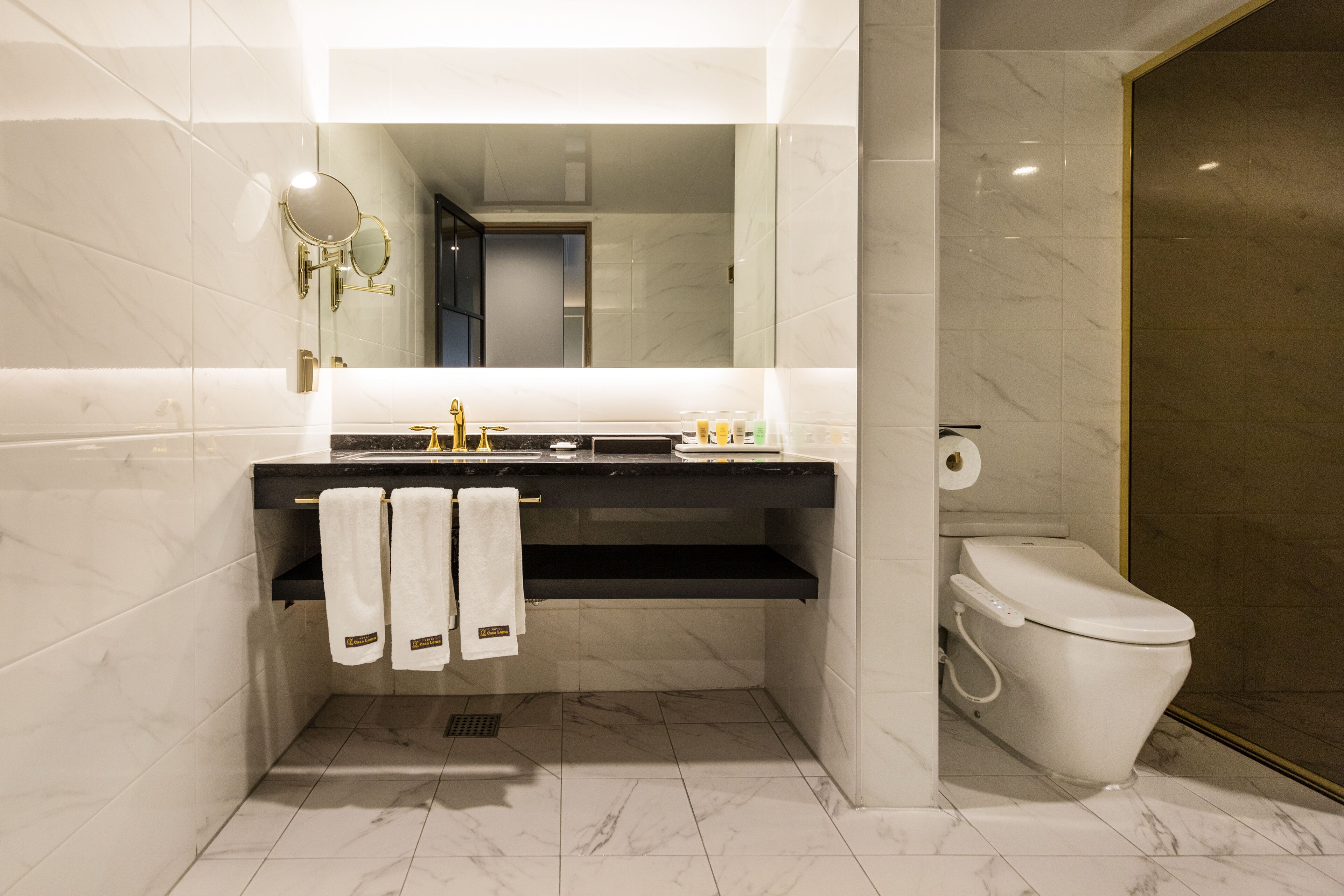 A modern bathroom with a black countertop, white tiles, a gold-framed mirror, and a toilet with a gold frame.