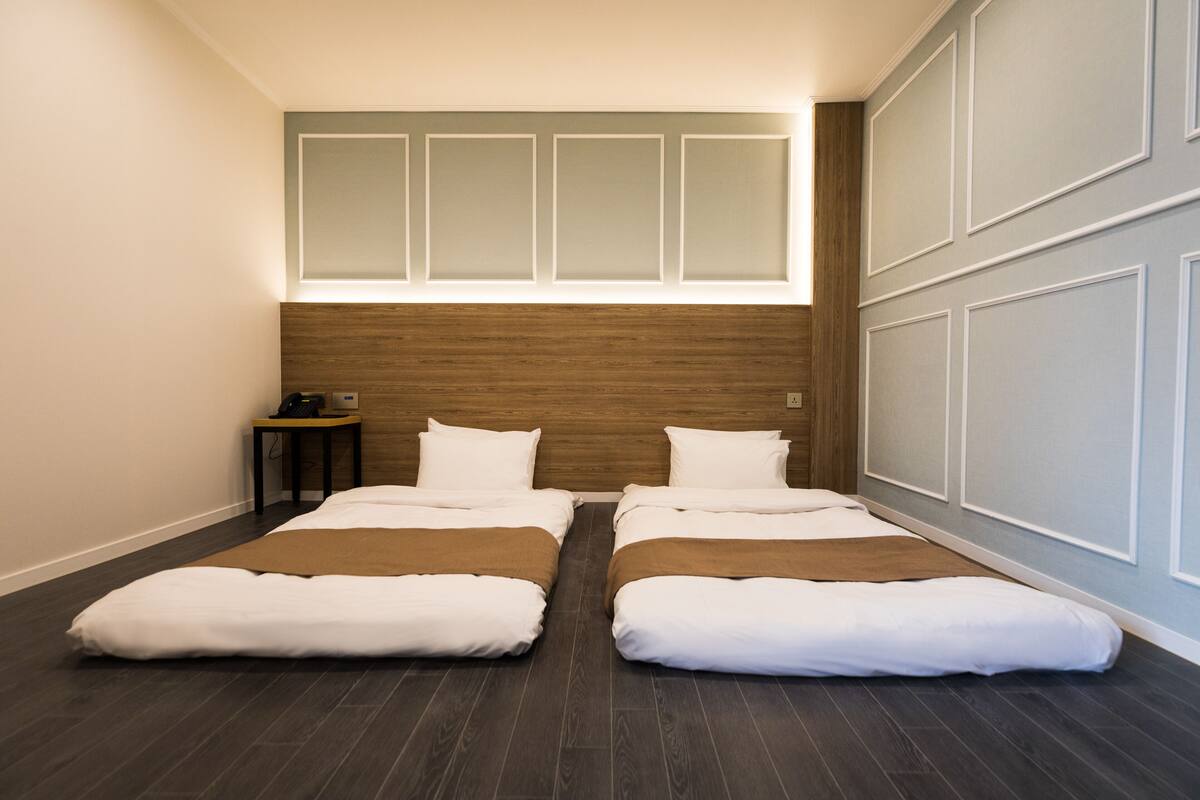 A hotel room with two beds, a wooden headboard, a small table with a lamp, and a wall-mounted shelf.
