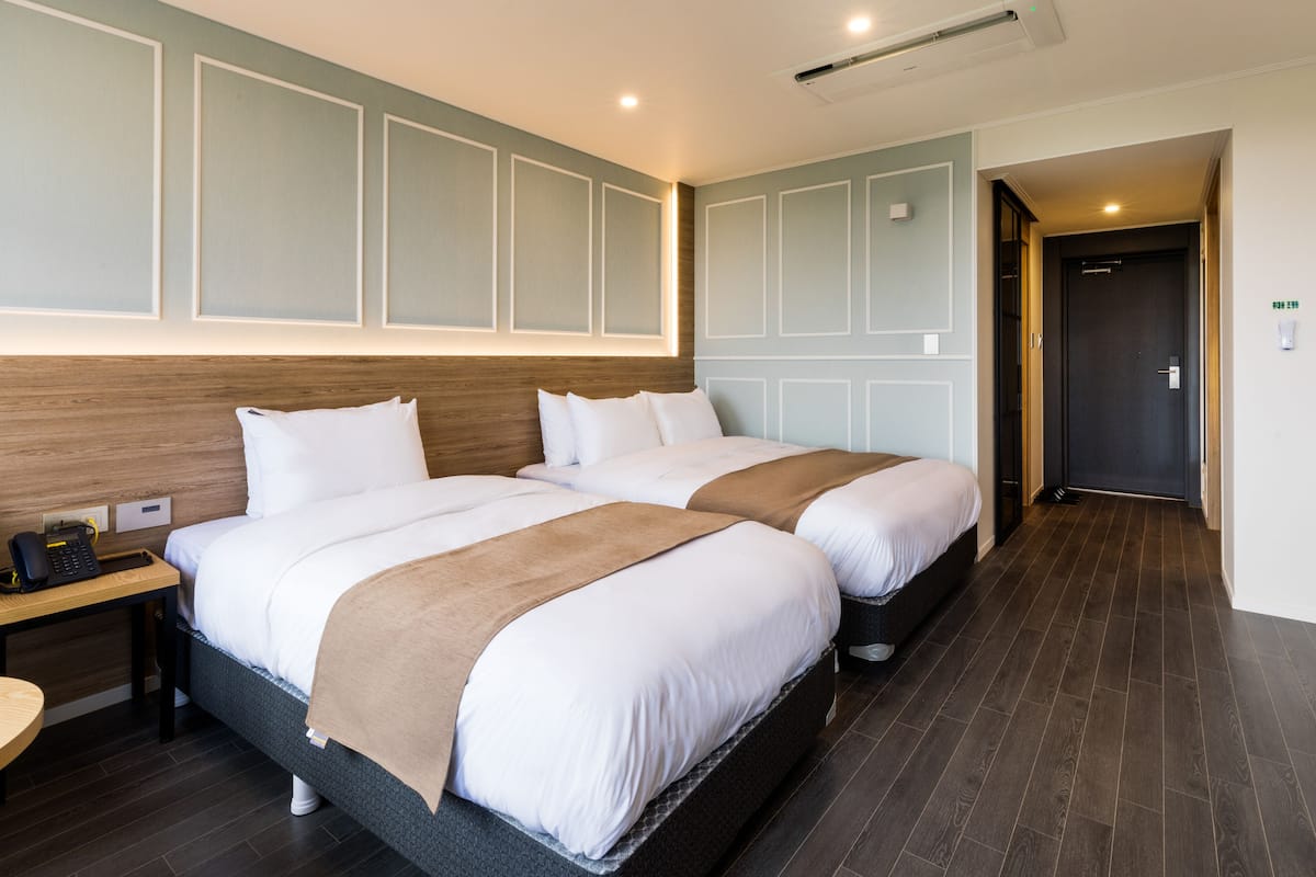 A hotel room with two beds, a wooden headboard, a bedside table with a phone, and a dark wooden floor.