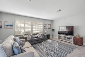 A modern living room with a grey sofa, a coffee table, a TV, and a large window with blinds. - Polynesian Gardens Villa: Renovated, Near Crescent Beach, Pool, Patio & Modern Comforts! (Siesta Key)