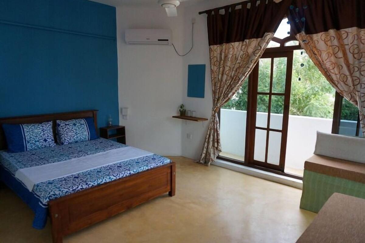 A bedroom with a wooden bed, a blue and white patterned bedspread, a small bedside table, a wall-mounted air conditioner, and a window with curtains.