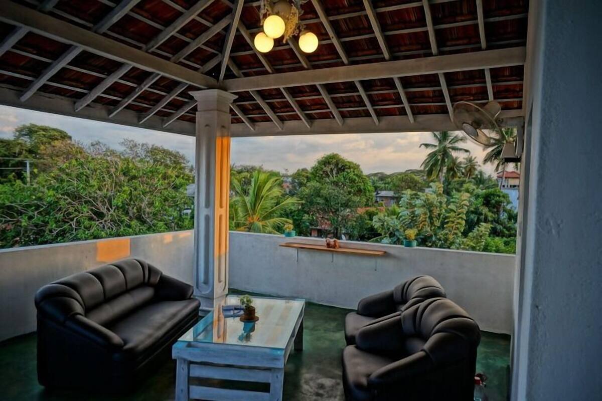 A covered outdoor seating area with two sofas and a small table, overlooking a lush garden with various plants and trees.