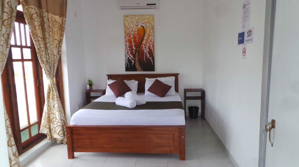 Un dormitorio con cama de madera, dos almohadas, una colcha, un aire acondicionado de pared, un cuadro y una puerta con un letrero.