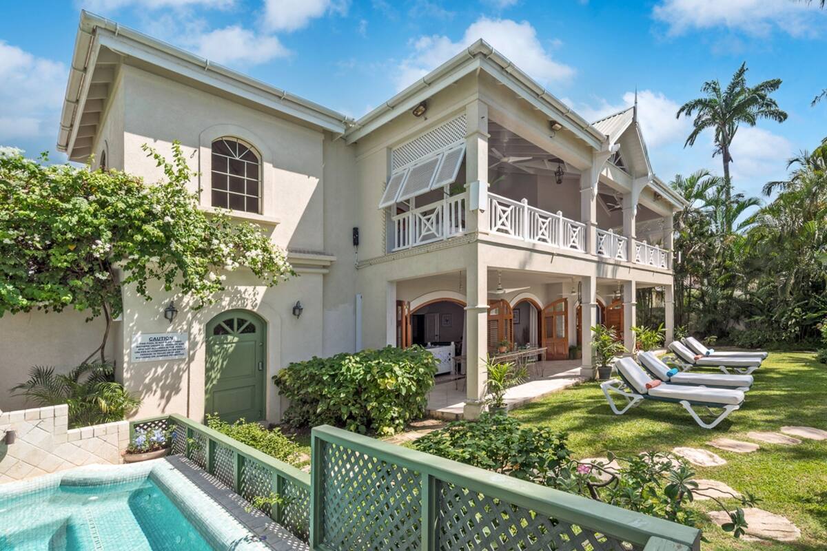 Um edifício de dois andares com piscina, alpendre coberto e jardim com cadeiras de lounge.