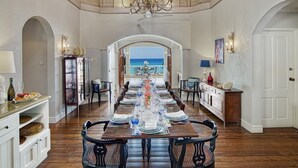 Comedor con una mesa larga dispuesta para una comida, suelo de madera y vistas al océano.