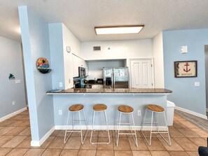 A kitchen with a central island, four bar stools, a refrigerator, and a microwave.