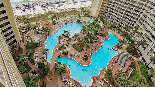 A hotel with a large, irregularly shaped pool surrounded by palm trees and lounge chairs. A beach is visible in the background. - 1322 Pool & Beach views, w/2 parking passes, lagoon-style pool & indoor pool (Panama City Beach)