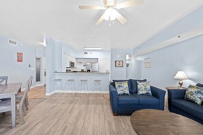 A modern living room with a blue sofa, wooden coffee table, and a dining area with a wooden table and chairs. - 1322 Pool & Beach views, w/2 parking passes, lagoon-style pool & indoor pool (Panama City Beach)