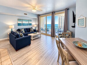 A living room with a sofa, coffee table, and dining area with chairs. There is a balcony with a view of the ocean and a building. - 1322 Pool & Beach views, w/2 parking passes, lagoon-style pool & indoor pool (Panama City Beach)