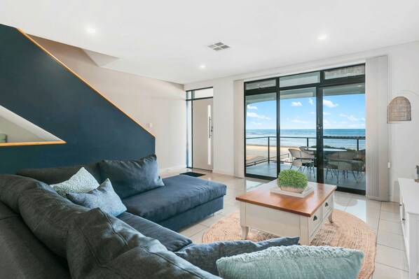 A modern living room with a dark-colored sofa, a wooden coffee table, and a view of the beach through large glass doors.
