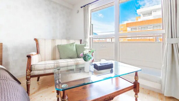 A room with a glass-top coffee table, a striped sofa with a green pillow, and a view of a building through a window. - Commence5 (Sapporo)
