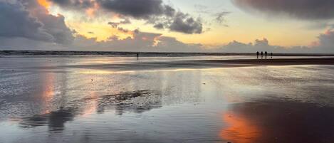 Una playa con arena mojada que refleja el cielo, algunas personas caminando y un atardecer al fondo.
