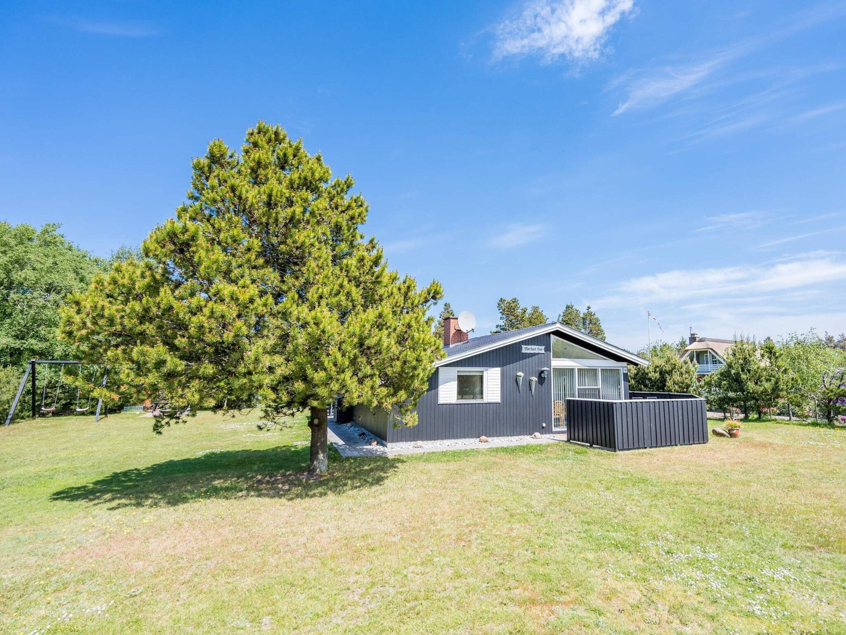 A single-story house with a large tree in the yard, a swing set, and a fenced area.