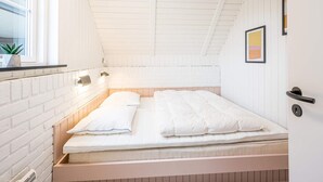 A small, white-walled room with a sloped ceiling, a single bed with white bedding, a window with blinds, a wall-mounted light, and a small potted plant. - 6 Person Holiday Home in Hvide Sande (Hvide Sande)