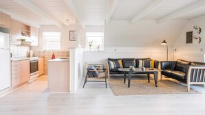 A spacious living area with a black leather sofa, a coffee table, and a dining table. There is a kitchen area with wooden cabinets and a stove. The room has white walls and a high ceiling with exposed beams. - 6 Person Holiday Home in Hvide Sande (Hvide Sande)