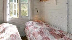 A bedroom with two beds, a window with curtains, a bedside lamp, and a shelf with books.