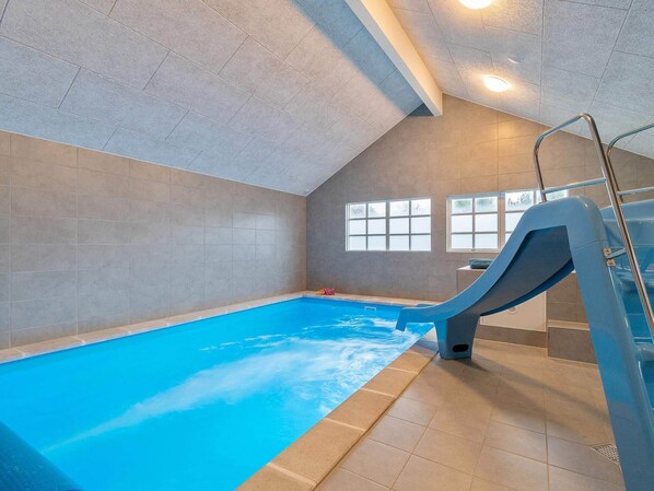 An indoor swimming pool with a slide, tiled walls, and windows.
