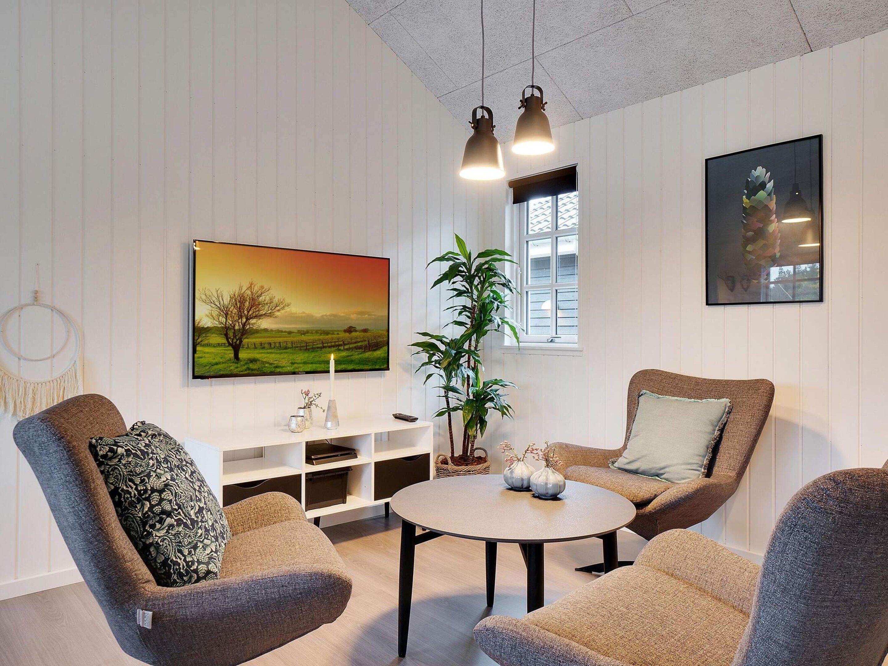 A living room with a round table, two armchairs, a large flat-screen TV, a potted plant, and a framed picture on the wall.