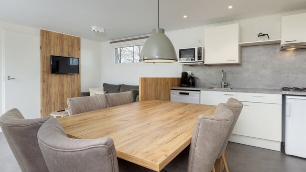 A modern kitchen with a wooden dining table, grey upholstered chairs, a built-in microwave, and a coffee maker. - Modern Chalet Near Forest (Gasselternijveen)