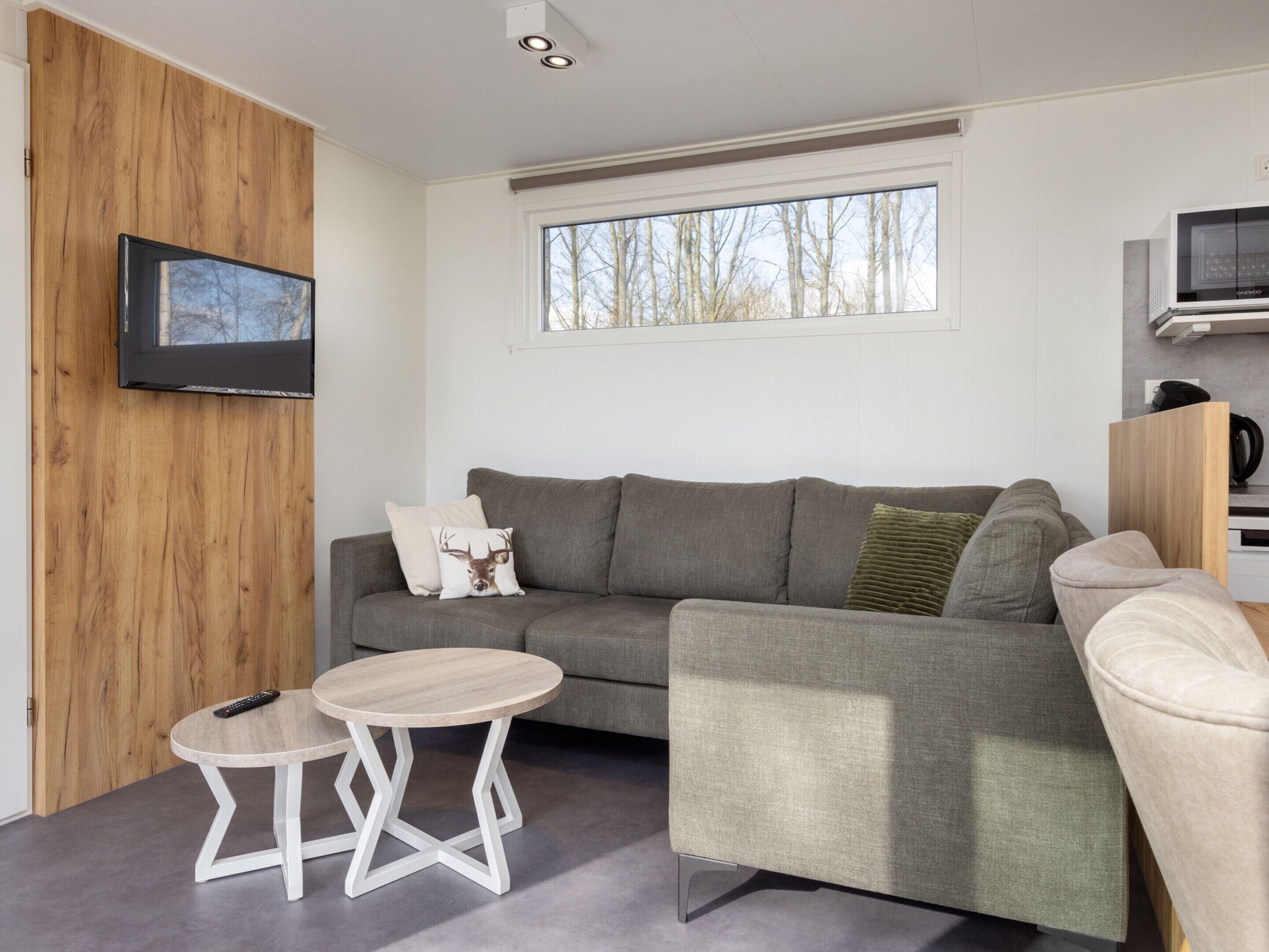 Een moderne woonkamer met een grijze bank, een klein rond tafeltje en een aan de muur bevestigde televisie.