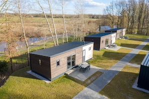 A modern building with a flat roof, a patio with outdoor furniture, and a view of a body of water and trees. - Modern Chalet Near Forest (Gasselternijveen)