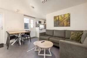A modern living room with a sofa, a dining table with chairs, a kitchen area with a microwave, and a large window. - Cozy Chalet w/ Pool Access (Gasselternijveen)