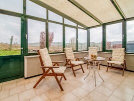 Un invernadero con cuatro sillas acolchadas y una mesita pequeña, ventanas amplias y vista a un campo.