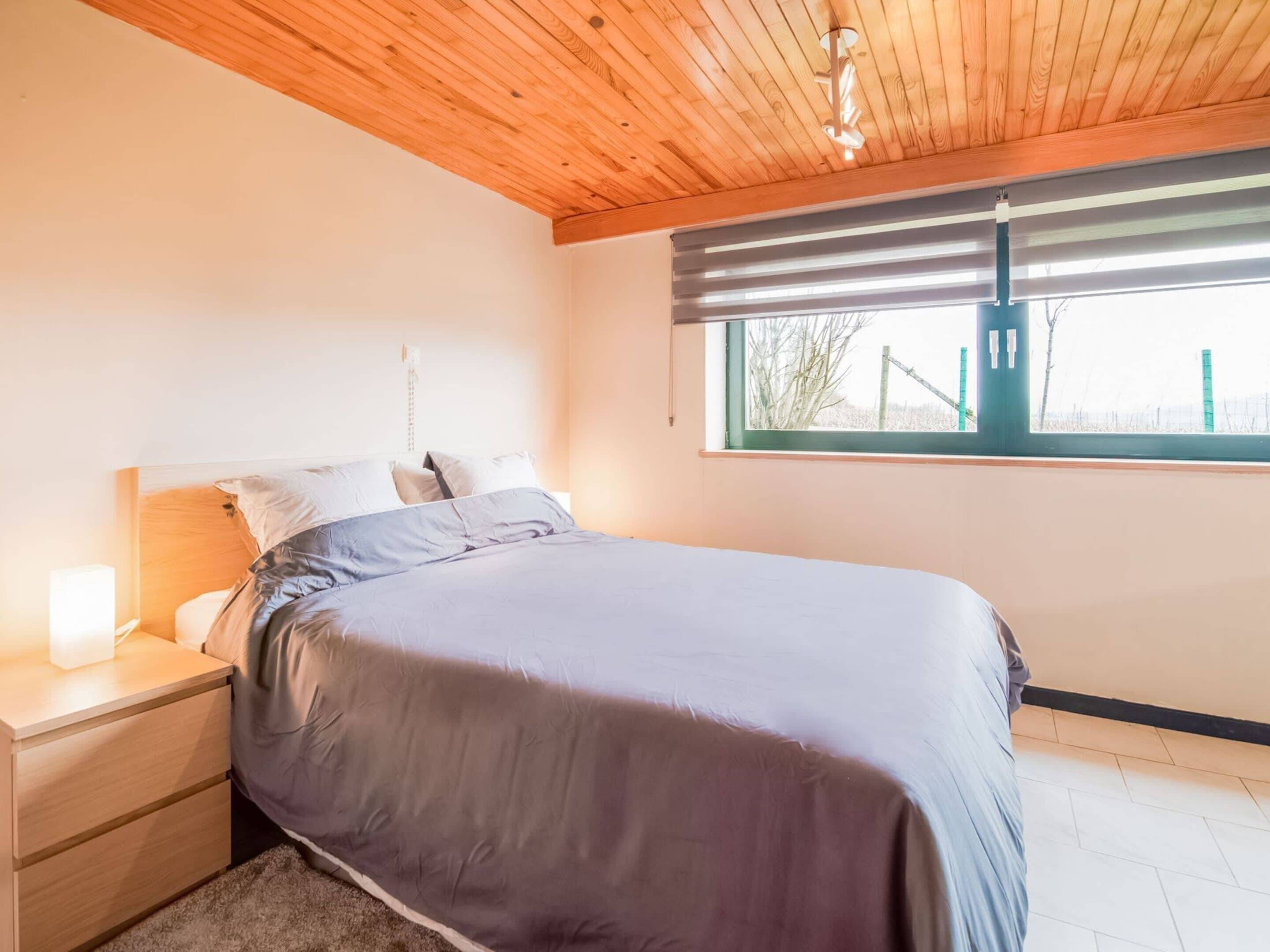 Een slaapkamer met een houten plafond, een bed met een grijs en wit dekbed, een nachtkastje met een lamp en een raam met jaloezieën.