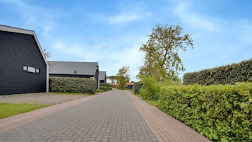 A paved pathway leading to modern buildings with dark siding and sloped roofs, surrounded by well-maintained hedges and trees.