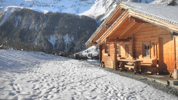 Uma cabana de madeira com varanda e uma mesa, rodeada por montanhas e árvores cobertas de neve.