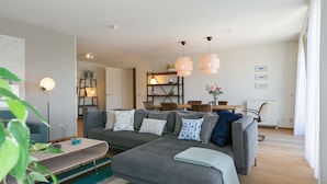 A modern living room with a grey sectional sofa, a wooden coffee table, and a striped rug. There is a dining area with a wooden table and chairs, and a bookshelf with decorative items. - Apartment in Den Haag With sea Beach (The Hague)