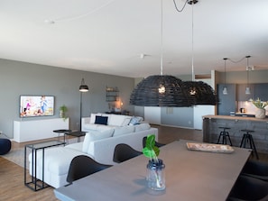 A modern living room with a sofa, a dining table, and a kitchen area in the background. - Apartment in Den Haag for 5 Person (The Hague)