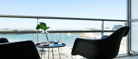 Un balcon avec deux chaises noires, une petite table ronde sur laquelle se trouvent un vase en verre et une plante, et une vue sur la mer et les bâtiments.