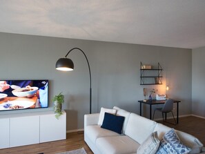 A modern living room with a flat-screen TV, a white sofa, a floor lamp, and a small table with a chair. - Apartment in Den Haag for 5 Person (The Hague)
