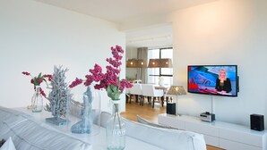 Una sala de estar moderna con un sofá blanco, un comedor con mesa y sillas, un televisor montado en la pared y jarrones decorativos con flores.