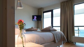 Un dormitorio con cama, televisión montada en la pared, ventana con cortinas y mesita de noche con un florero.
