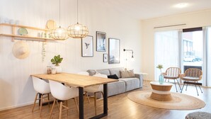 A modern living room with a wooden dining table, white chairs, a grey sofa, wicker chairs, and a large window with a view of the street. - Ground Floor Apartment near Scheveningen (The Hague)